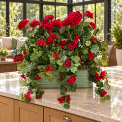 Three artificial red geraniums in a sage green metal planter on an enclosed patio countertop