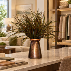 21-inch artificial sage green and brown grass arrangement in a riveted copper-tone container on an interior-design showroom counter.