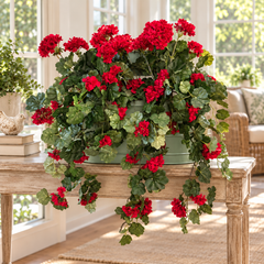 Three artificial red geraniums in a sage green metal planter on a sunroom console table