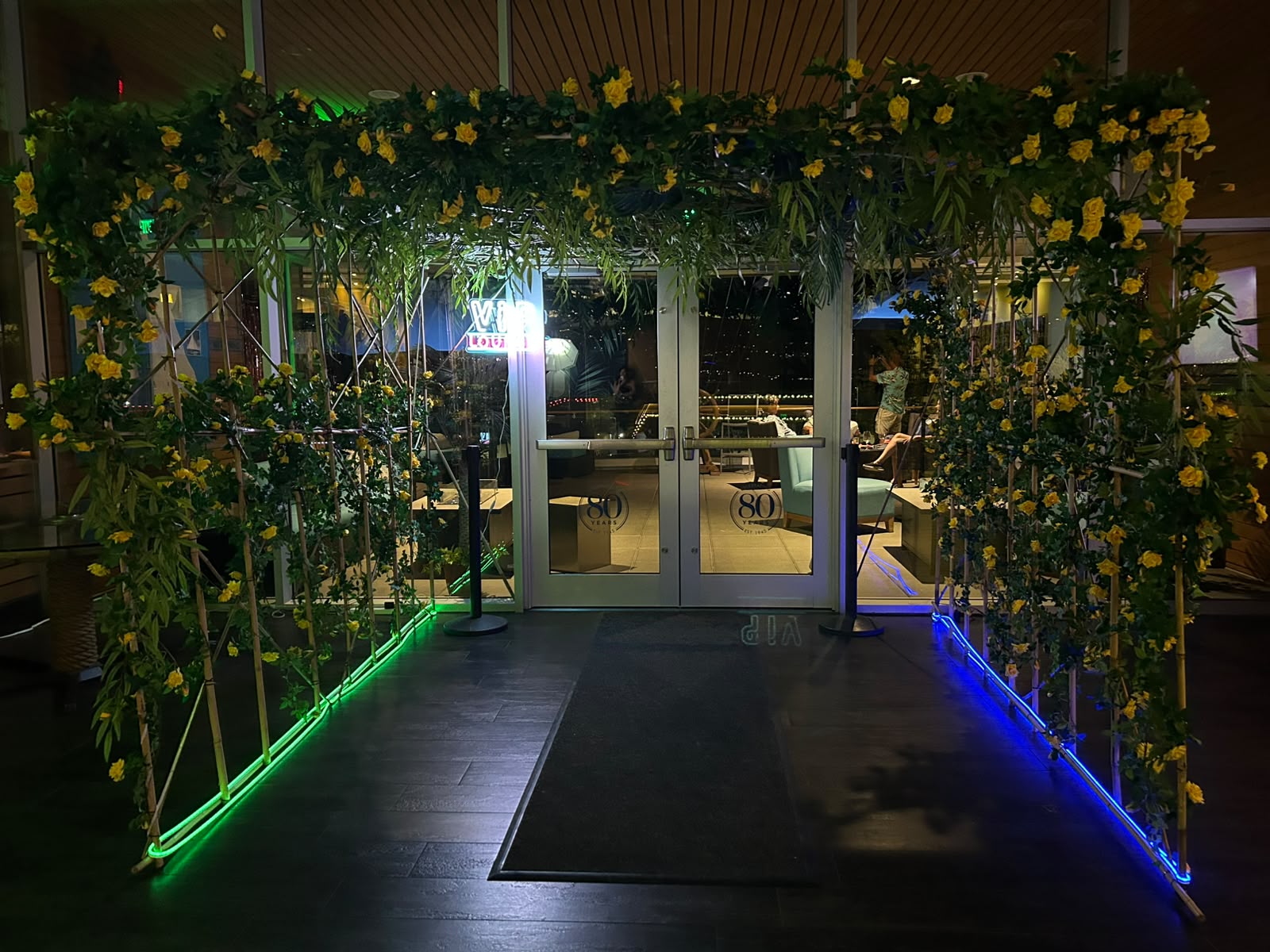 A decorative entrance archway with artificial rose bush vines, weeping willow, salloum leaves and is illuminated by green and blue LED strip lights along the base, set in front of glass doors.