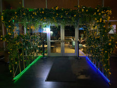 A decorative entrance archway with artificial rose bush vines, weeping willow, salloum leaves and is illuminated by green and blue LED strip lights along the base, set in front of glass doors.
